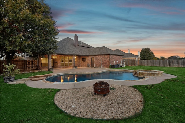 pool at dusk with a yard, an in ground hot tub, central AC unit, a patio area, and an outdoor fire pit