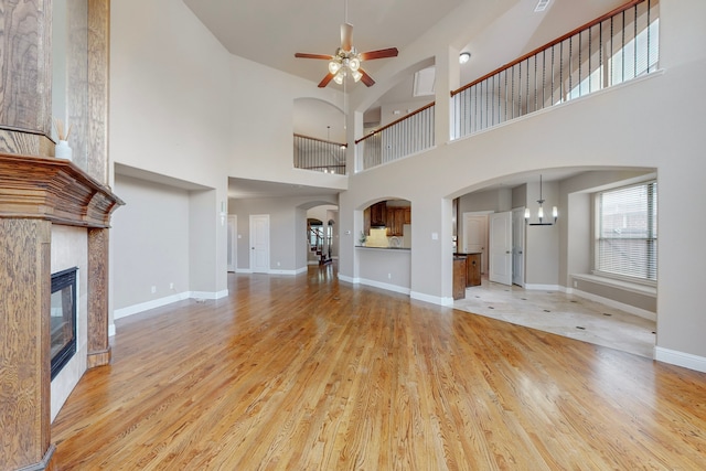 unfurnished living room with a tile fireplace, a towering ceiling, light hardwood / wood-style floors, and ceiling fan with notable chandelier