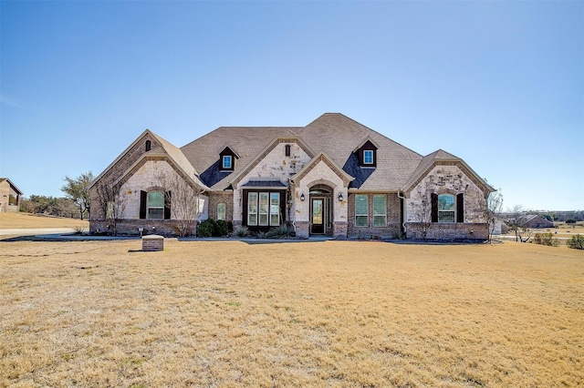 french country inspired facade with a shingled roof, stone siding, brick siding, and a front lawn