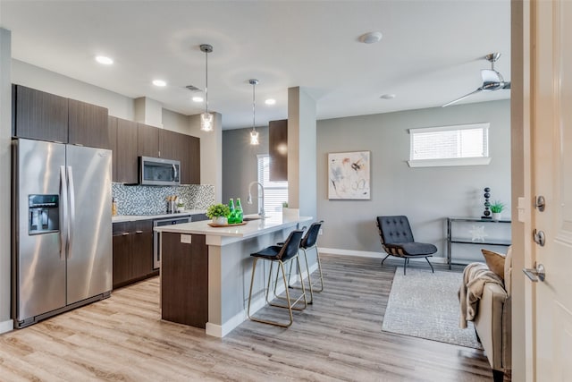 kitchen with dark brown cabinetry, stainless steel appliances, pendant lighting, light hardwood / wood-style floors, and a kitchen island with sink