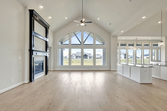 unfurnished living room with ceiling fan with notable chandelier, light hardwood / wood-style floors, high vaulted ceiling, and a wealth of natural light