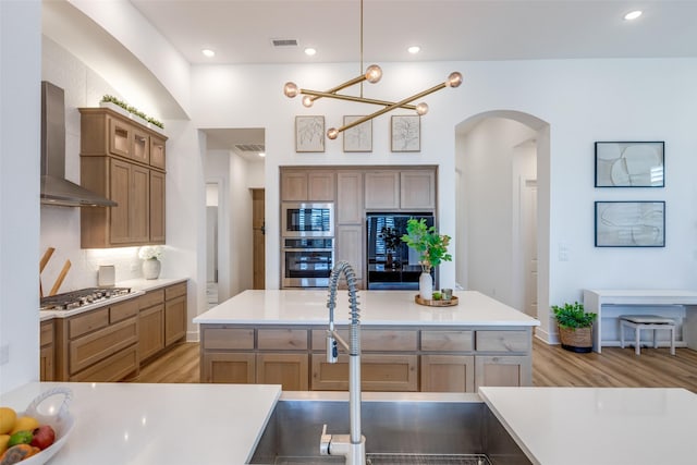 kitchen featuring a kitchen island with sink, wall chimney exhaust hood, decorative light fixtures, stainless steel appliances, and a chandelier