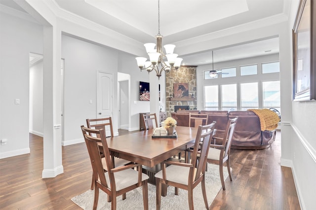 dining room featuring ceiling fan with notable chandelier, a stone fireplace, a raised ceiling, crown molding, and dark hardwood / wood-style floors