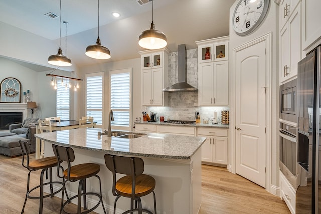 kitchen featuring light stone countertops, wall chimney exhaust hood, an island with sink, pendant lighting, and white cabinets