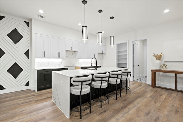 kitchen with sink, hanging light fixtures, backsplash, an island with sink, and white cabinets