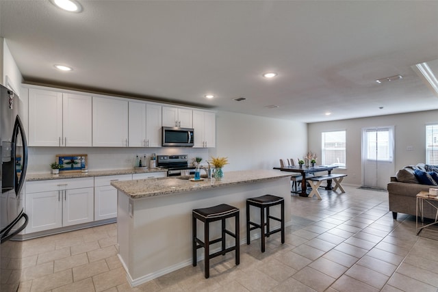 kitchen with a kitchen breakfast bar, light stone counters, stainless steel appliances, a kitchen island with sink, and white cabinetry