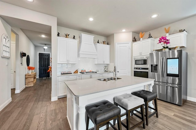 kitchen featuring custom exhaust hood, white cabinets, sink, an island with sink, and appliances with stainless steel finishes