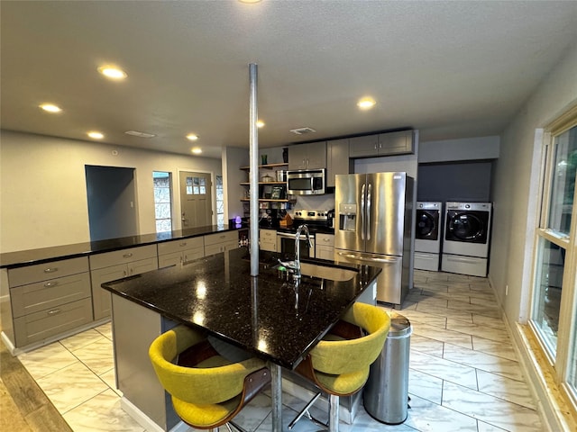 kitchen with gray cabinetry, stainless steel appliances, dark stone countertops, washer and clothes dryer, and a center island with sink