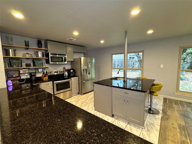 kitchen with sink, gray cabinets, appliances with stainless steel finishes, a kitchen island with sink, and dark stone counters