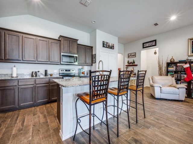 kitchen featuring a breakfast bar area, dark brown cabinets, stainless steel appliances, light stone countertops, and a center island with sink