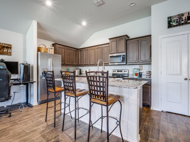 kitchen featuring appliances with stainless steel finishes, wood-type flooring, a kitchen bar, a kitchen island with sink, and light stone counters