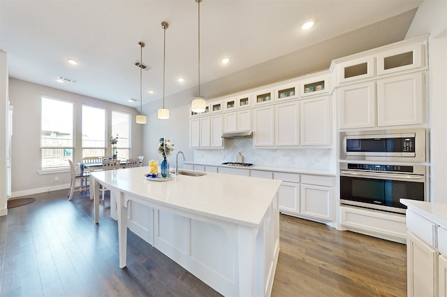 kitchen featuring backsplash, white cabinets, an island with sink, appliances with stainless steel finishes, and decorative light fixtures