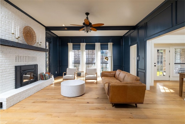 living room with a brick fireplace, light wood-type flooring, beam ceiling, ornamental molding, and ceiling fan