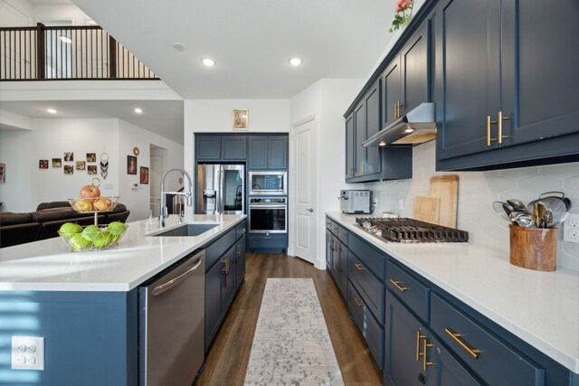 kitchen featuring dark wood-type flooring, range hood, blue cabinets, a center island with sink, and appliances with stainless steel finishes