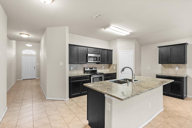 kitchen with appliances with stainless steel finishes, sink, backsplash, a kitchen island with sink, and light stone countertops