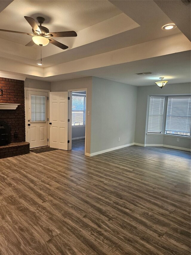 unfurnished living room featuring a tray ceiling, ceiling fan, dark hardwood / wood-style flooring, and a brick fireplace