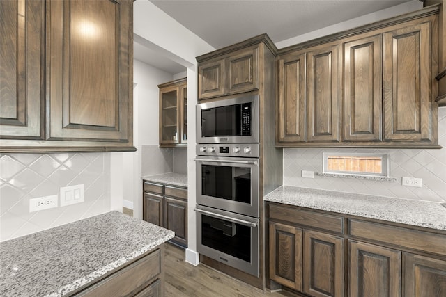 kitchen with stainless steel double oven, light stone countertops, backsplash, built in microwave, and glass insert cabinets