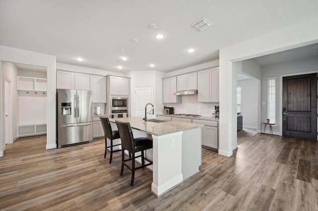 kitchen with sink, stainless steel appliances, light stone counters, a center island with sink, and hardwood / wood-style flooring