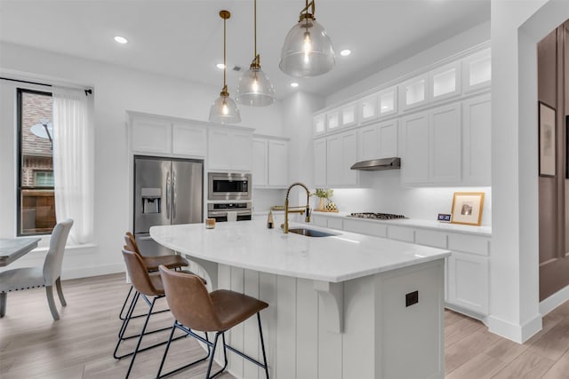 kitchen featuring a kitchen island with sink, stainless steel appliances, a sink, white cabinetry, and decorative light fixtures