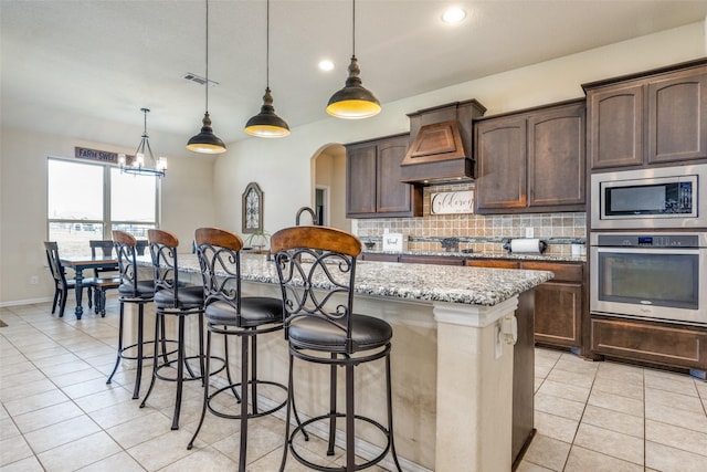 kitchen featuring a kitchen island with sink, hanging light fixtures, light stone countertops, appliances with stainless steel finishes, and dark brown cabinets