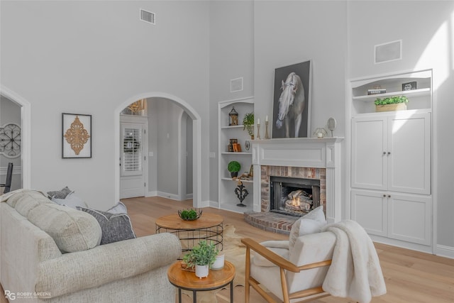 living room featuring a brick fireplace, a towering ceiling, built in features, and light wood-type flooring