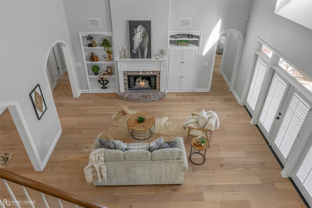 living room featuring a brick fireplace, built in shelves, a towering ceiling, and light wood-type flooring