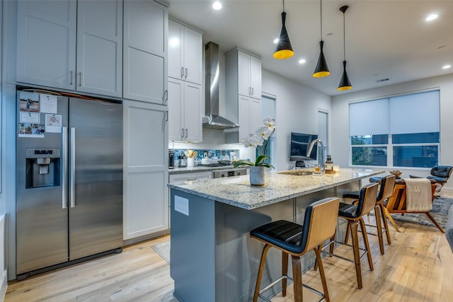 kitchen with light stone countertops, stainless steel appliances, wall chimney range hood, decorative light fixtures, and a center island with sink