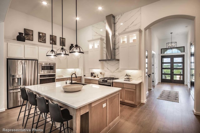 4121 Buffalo Clover Dr, Aubrey, TX 76227 - kitchen featuring appliances with stainless steel finishes, wall chimney exhaust hood, arched walkways, a towering ceiling, and a kitchen island with sink