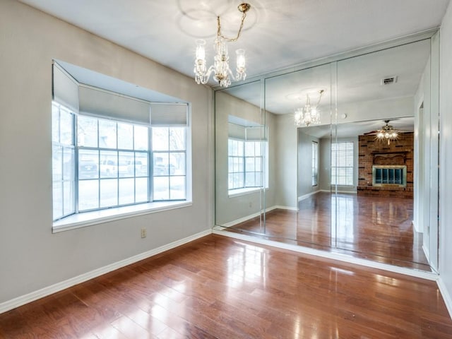 unfurnished dining area with ceiling fan with notable chandelier, a brick fireplace, and hardwood / wood-style floors