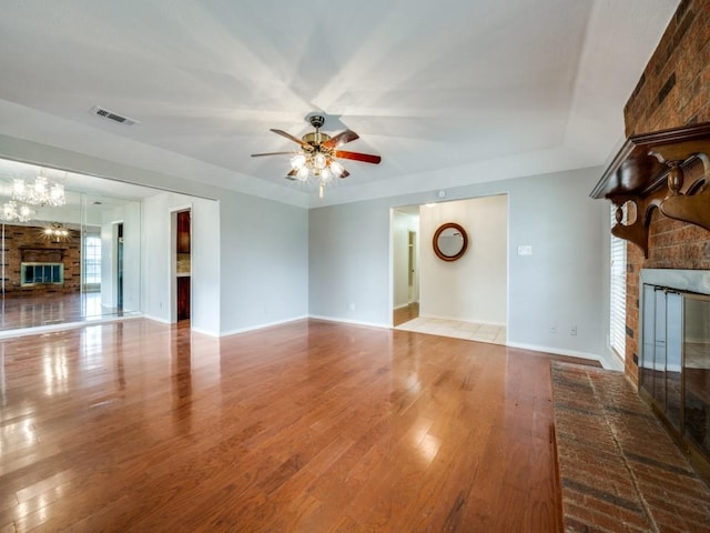 unfurnished living room with a brick fireplace, a tray ceiling, ceiling fan with notable chandelier, and light hardwood / wood-style floors