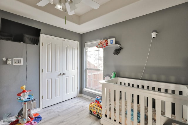 bedroom featuring a crib, ceiling fan, light hardwood / wood-style floors, a raised ceiling, and a closet