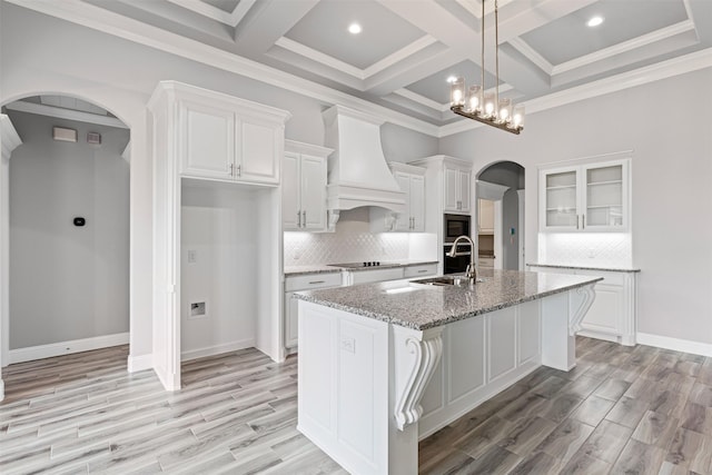 kitchen with light stone countertops, custom exhaust hood, a center island with sink, light hardwood / wood-style flooring, and white cabinets