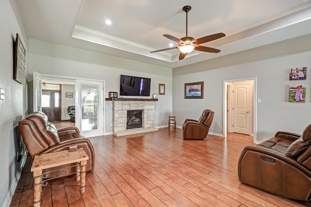 living room with a stone fireplace, light wood-type flooring, ornamental molding, a tray ceiling, and ceiling fan