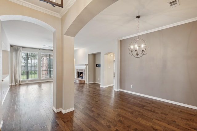 unfurnished room featuring crown molding, dark wood-type flooring, ceiling fan with notable chandelier, and a brick fireplace