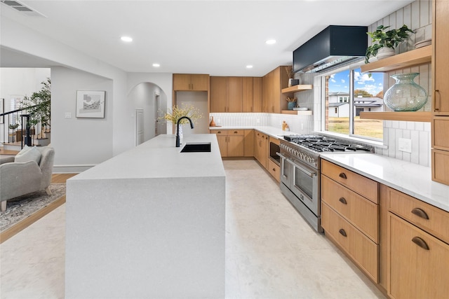 kitchen featuring sink, wall chimney range hood, an island with sink, range with two ovens, and decorative backsplash