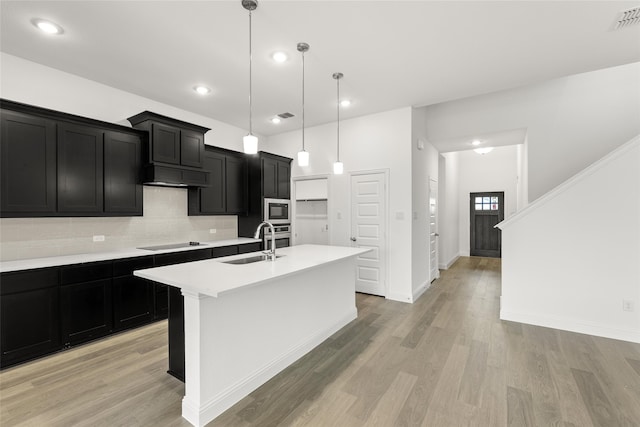 kitchen featuring black electric stovetop, sink, hanging light fixtures, a center island with sink, and built in microwave