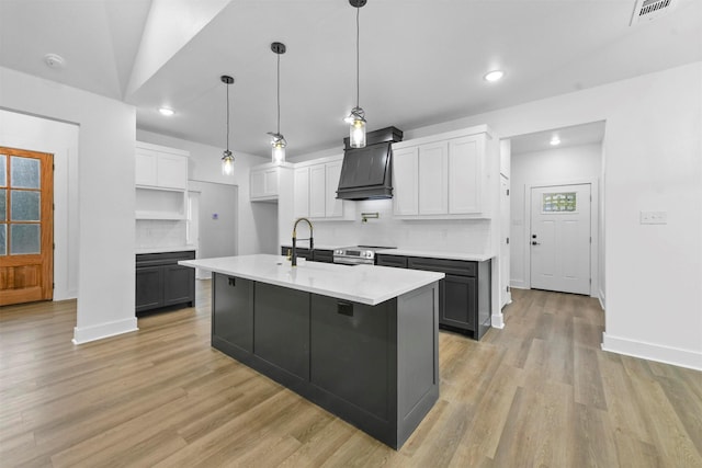 kitchen featuring backsplash, premium range hood, light hardwood / wood-style floors, white cabinetry, and an island with sink