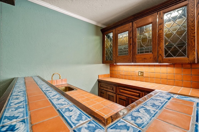 bar featuring tasteful backsplash, sink, tile counters, crown molding, and a textured ceiling