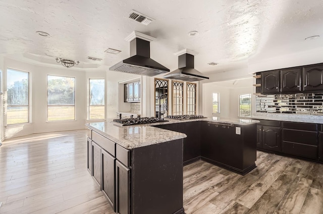 kitchen featuring wall chimney range hood, light hardwood / wood-style flooring, island exhaust hood, stainless steel gas stovetop, and a kitchen island