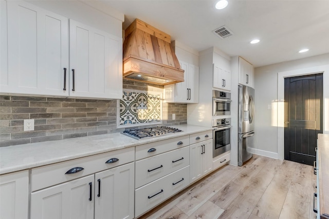 kitchen with premium range hood, appliances with stainless steel finishes, white cabinets, backsplash, and light hardwood / wood-style flooring