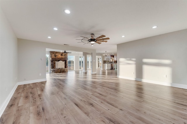 unfurnished living room with a stone fireplace, ceiling fan with notable chandelier, and light wood-type flooring