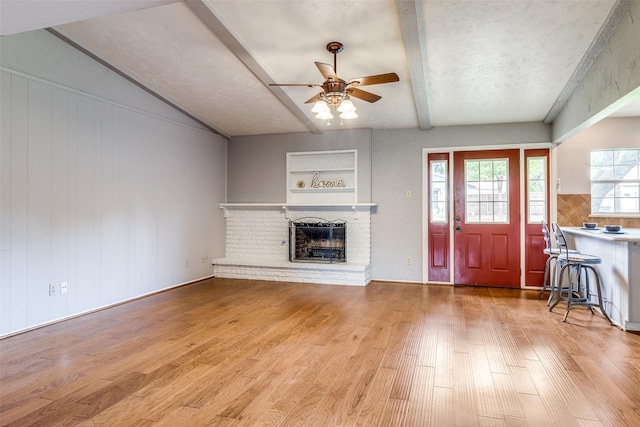 unfurnished living room featuring ceiling fan, beam ceiling, a brick fireplace, and light wood-type flooring