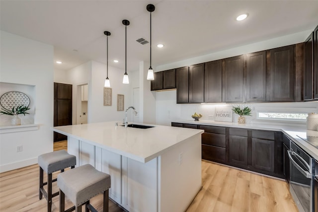 kitchen with stainless steel oven, a center island with sink, sink, decorative light fixtures, and light hardwood / wood-style floors