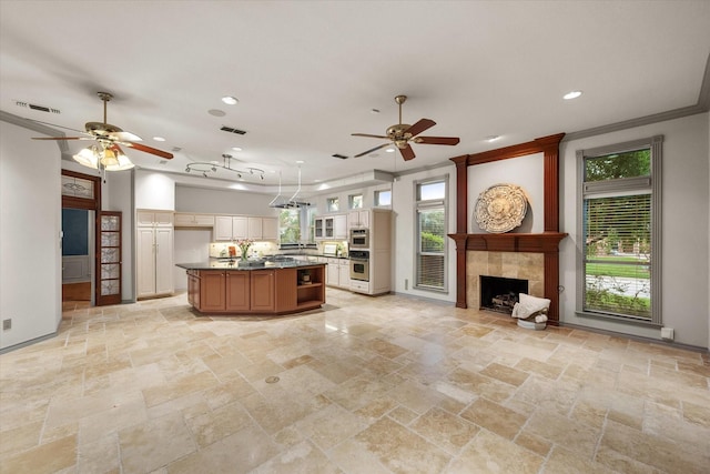 kitchen featuring crown molding, a tile fireplace, ceiling fan, a center island with sink, and stainless steel double oven