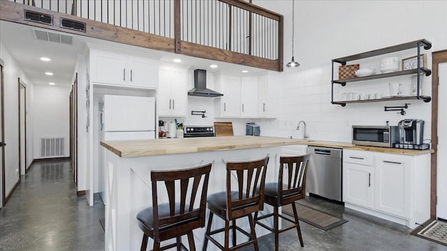 kitchen featuring butcher block counters, stainless steel appliances, wall chimney range hood, a kitchen bar, and white cabinets