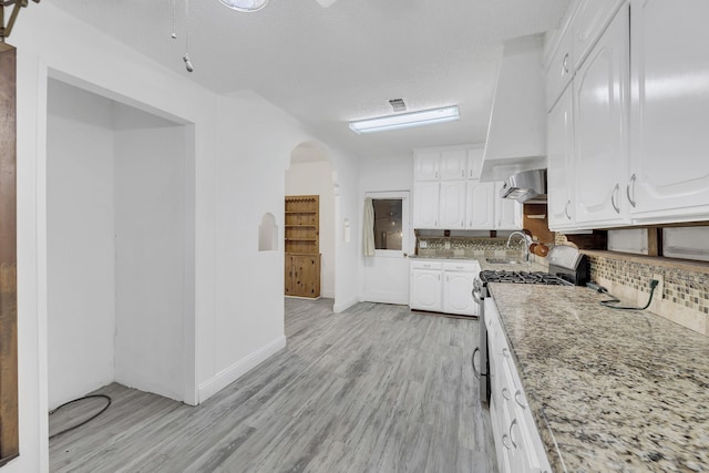 kitchen with wall chimney range hood, stainless steel gas stove, tasteful backsplash, light stone counters, and white cabinetry