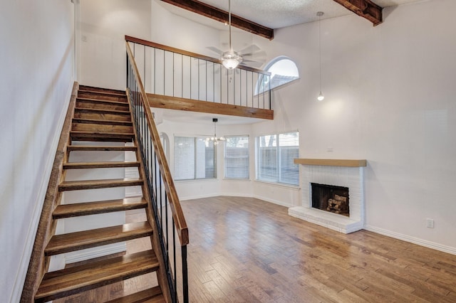 staircase with beam ceiling, a brick fireplace, a towering ceiling, wood-type flooring, and ceiling fan with notable chandelier