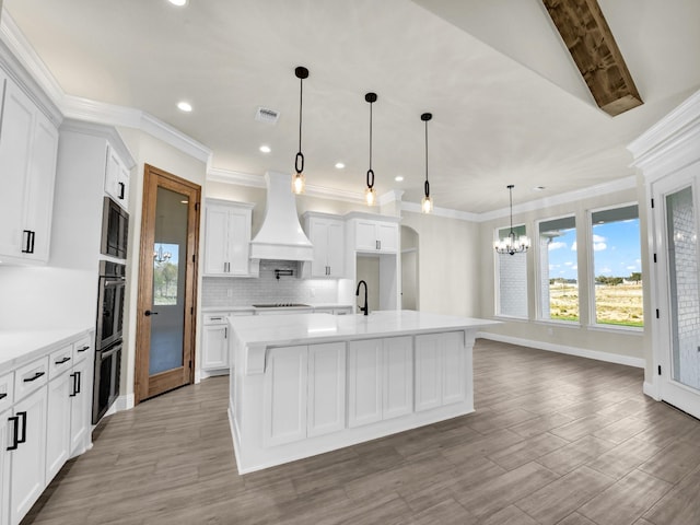 kitchen with custom exhaust hood, a kitchen island with sink, sink, decorative light fixtures, and white cabinetry