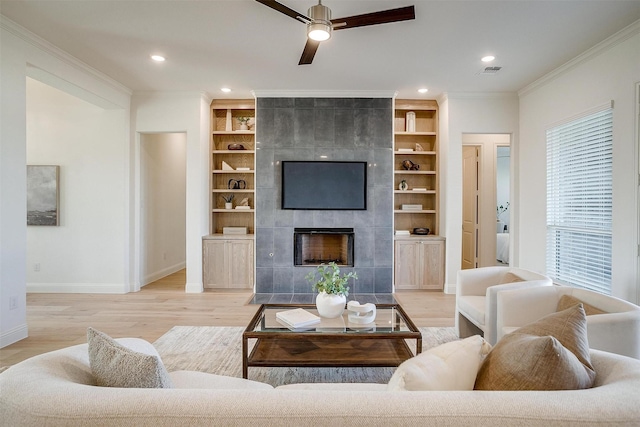 living room with ceiling fan, built in features, crown molding, a tiled fireplace, and light wood-type flooring