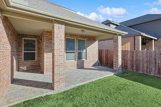 view of exterior entry featuring a shingled roof, a lawn, fence, a patio area, and brick siding